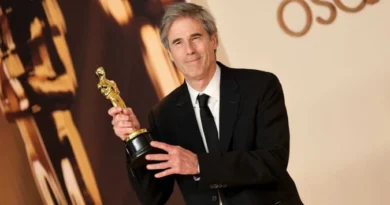 Homem de terno preto levanta estatueta do Oscar diante de painel com a palavra OSCARS e outro troféu desfocado ao fundo.