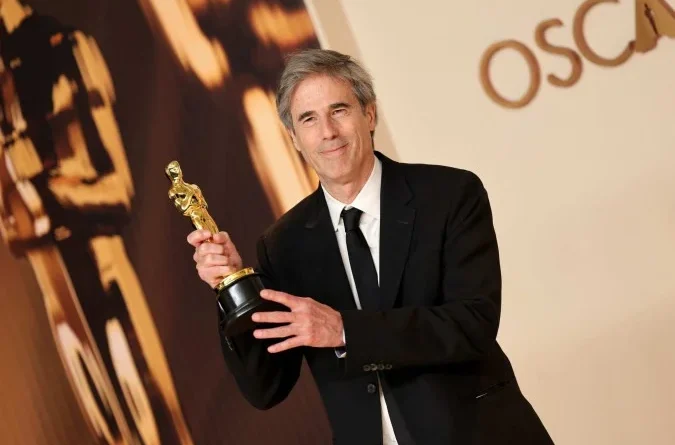 Homem de terno preto levanta estatueta do Oscar diante de painel com a palavra OSCARS e outro troféu desfocado ao fundo.