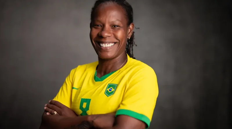 Pessoa sorrindo, de braços cruzados, veste camisa amarela da seleção brasileira diante de fundo escuro.