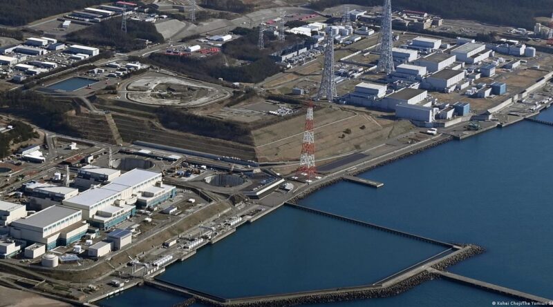 Vista aérea de usina nuclear no Japão, com prédios brancos, torres e tanques perto da costa, entre água e colinas verdes.