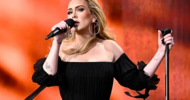 Mulher com longos cabelos loiros e vestido preto sem ombros canta no palco com fundo vermelho-alaranjado, segurando o pedestal do microfone.