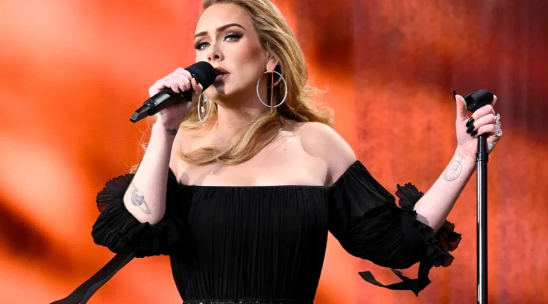 Mulher com longos cabelos loiros e vestido preto sem ombros canta no palco com fundo vermelho-alaranjado, segurando o pedestal do microfone.