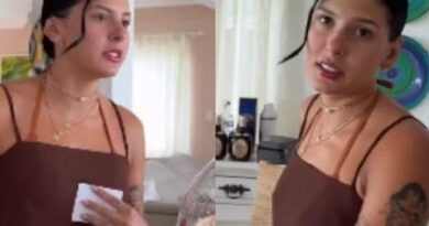 Mulher com cabelos escuros em um coque, vestido marrom sem mangas e colares segura um guardanapo em ambiente interno com tatuagens visíveis.