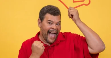 Homem sorridente de camisa vermelha segura o contorno de uma coroa acima da cabeça com uma mão e fecha o punho com a outra em um fundo amarelo.