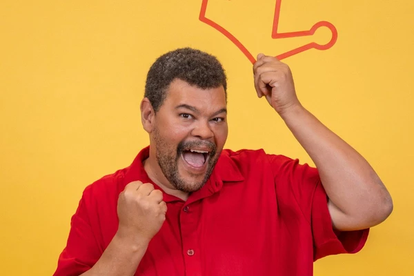 Homem sorridente de camisa vermelha segura o contorno de uma coroa acima da cabeça com uma mão e fecha o punho com a outra em um fundo amarelo.
