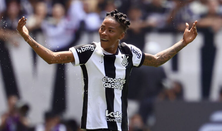 Jogador do Corinthians sorri com braços abertos no campo; torcida desfocada ao fundo em estádio de Londres.