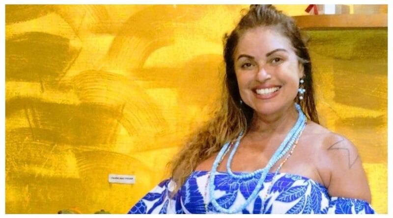 Mulher de cabelos castanhos sorri, usa vestido azul e branco e colar. Fundo amarelo destaca manchete sobre injúria racial.