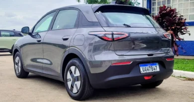Carro elétrico Geely EX2 cinza com placa brasileira estacionado próximo a um prédio azul e branco, com luzes traseiras arredondadas.
