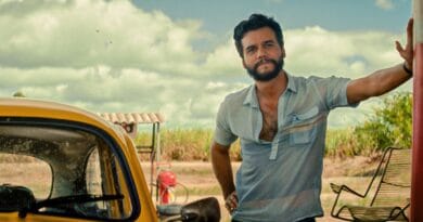 Homem de barba e camisa azul clara aberta ao lado de carro amarelo, com banco, grama e céu nublado ao fundo.