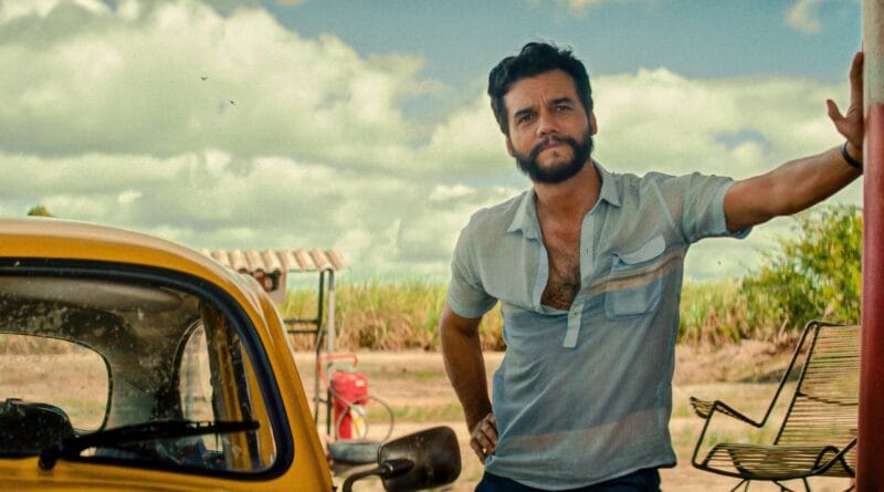 Homem de barba e camisa azul clara aberta ao lado de carro amarelo, com banco, grama e céu nublado ao fundo.