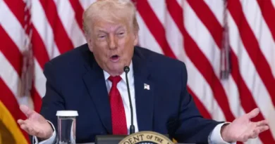 Homem de terno azul-marinho e gravata vermelha fala em uma mesa com microfone, selo presidencial de Trump, copo d'água e bandeiras dos EUA ao fundo.