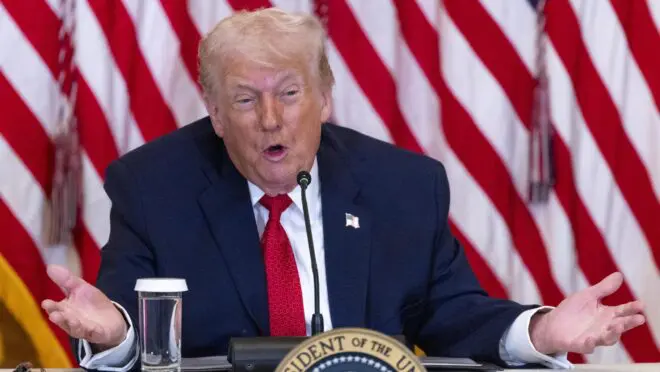 Homem de terno azul-marinho e gravata vermelha fala em uma mesa com microfone, selo presidencial de Trump, copo d'água e bandeiras dos EUA ao fundo.