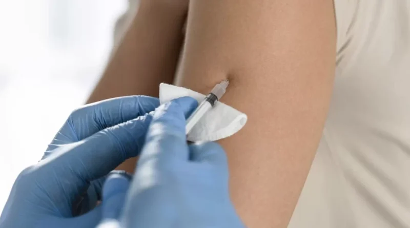 Profissional com luvas azuis aplica vacina contra câncer de pele no braço de paciente, segurando o olhar sobre a pele.